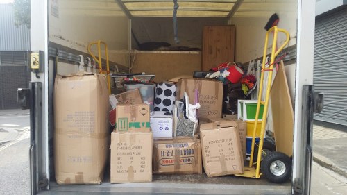 The image shows the interior of a mobile waste collection container, packed with various types of rubbish including large cardboard boxes, some partially crushed, with printed labels and barcodes, and a few smaller boxes stacked on top. To the left, a sizeable brown paper bag, crumpled and leaning against the boxes, is visible. On the right side, two yellow hand trucks are positioned, each with a handle extending upward, one with a small black item resting on top. A wooden cabinet or panel is situated at the back of the container, partially obstructed by the waste items. The surrounding environment suggests an industrial or commercial area, with part of a metallic roller shutter and some pavement visible outside. The scene is well-lit, likely during daylight, emphasizing the textures and colors of the waste materials, which include various shades of brown, beige, and black, with some red and green accents from packaging or other objects. This image aligns with the services offered by Business Waste Removal Wood Green, demonstrating typical rubbish often handled in local waste removal operations in the area.