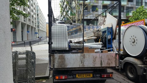 A small flatbed truck parked on a city street corner, loaded with various types of rubbish including white plastic items, metallic debris, and wooden waste. The truck's cargo area is filled to capacity with some waste stacked and leaning slightly to one side. The vehicle has a rusty, weathered appearance with a yellow license plate reading 'NX12 GHA'. It is positioned near a modern, multi-story building with balconies and large windows, and the street around it appears quiet with minimal traffic. In the background, there is a traffic light, some street signs, and a pedestrian crossing. The scene suggests a waste removal service likely operating in an urban area of London, possibly near Wood Green, aligned with local rubbish clearance for businesses or residents. The image conveys an active waste collection operation, typical of services provided by Business Waste Removal Wood Green.