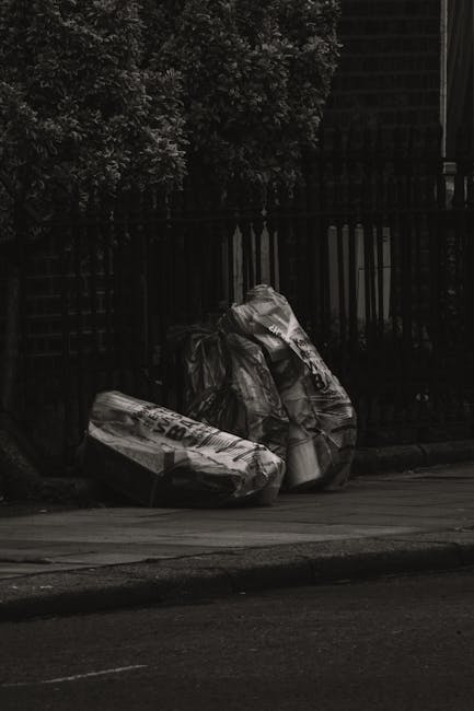Two large black plastic bags filled with unknown waste material rest on a paved sidewalk near a black metal fence, with a dense leafy hedge in the background. The bags appear to be tightly closed and