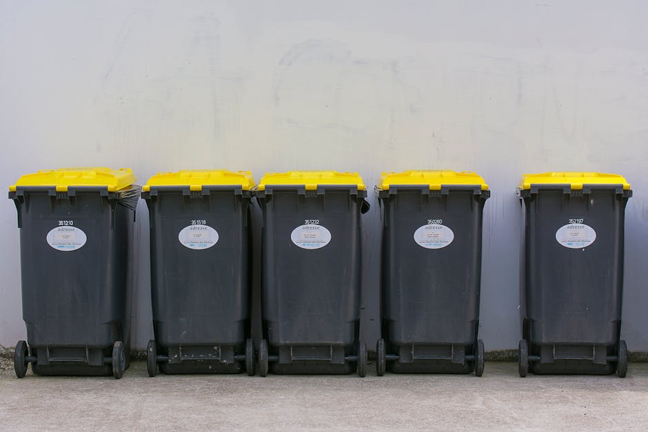 Five large black wheelie bins with yellow lids lined up against a plain light grey wall, positioned on a paved surface. Each bin has a white label with identification numbers and a barcode, indicating