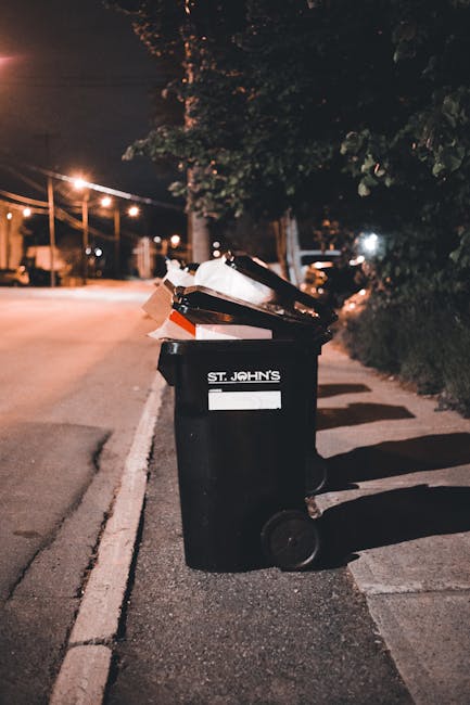 Five large black wheelie bins with yellow lids lined up against a plain light grey wall, positioned on a paved surface. Each bin has a white label with identification numbers and a barcode, indicating