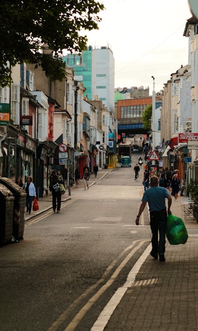 A city street scene showing a small pedestrianized area with a mix of commercial shops and cafes on either side, featuring modern and older architectural styles with some buildings painted in neutral 