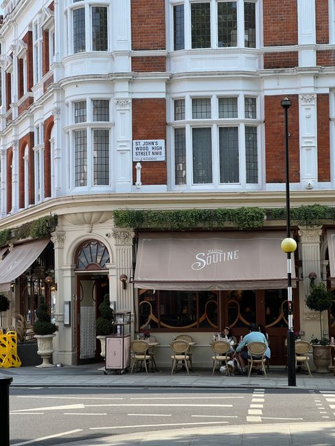 A city street scene showing a small pedestrianized area with a mix of commercial shops and cafes on either side, featuring modern and older architectural styles with some buildings painted in neutral 